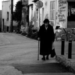 Black and white image of an elderly woman walking with a cane on a graffiti-lined urban street.